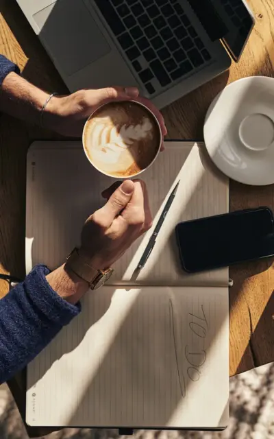 Person hält eine Tasse Kaffee und sitzt an einem Tisch mit Laptop.