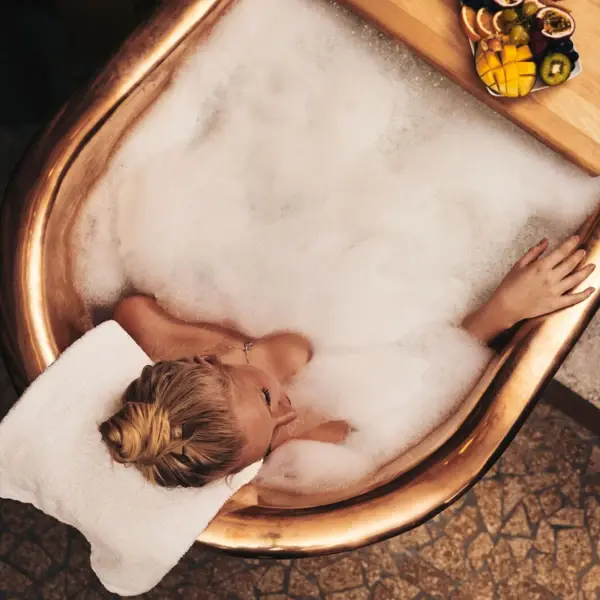 Eine Frau liegt in einer bronzefarbenen Badewanne, die mit viel Schaum gefüllt ist und entspannt. 
