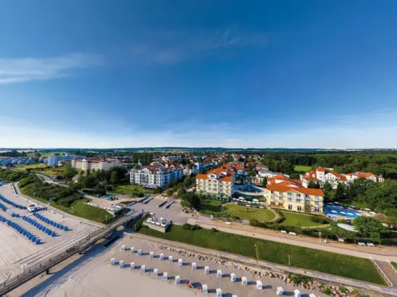 Luftaufnahme eines Hotelkomplexes am Strand vor einer Stadt mit Gebäuden, Bäumen und Wolken.