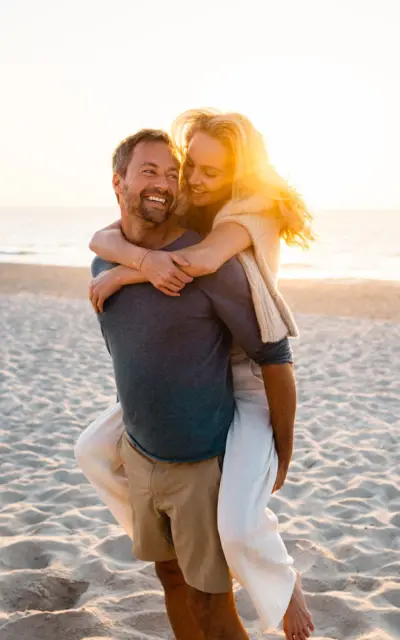 Mann trägt eine Frau auf seinem Rücken am Strand, beide lächeln und umarmen sich.