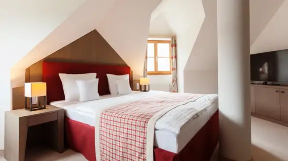 Ein rotes Polsterbett steht zwischen einer Wand und einer Säule und es liegt eine karierte Tagesdecke am Fußende. Es stehen jeweils eine Lampe links und rechts neben dem Bett auf Nachtschränken. Im Hintergrund ist ein kleines Fenster zu sehen.