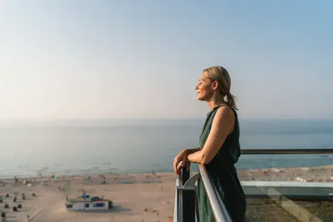 Frau in Kleidung steht auf einem Balkon und blickt auf einen Strand.