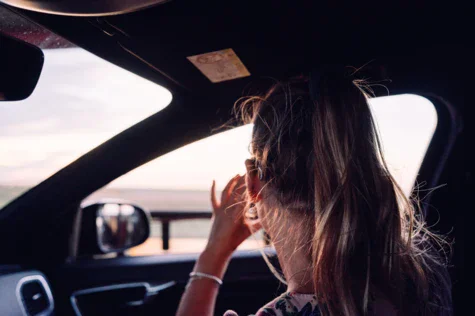 Eine Beifahrerin ist von hinten fotografiert. Sie schaut aus dem Fenster auf das den Sonnenuntergang widerspiegelndes Meer. 