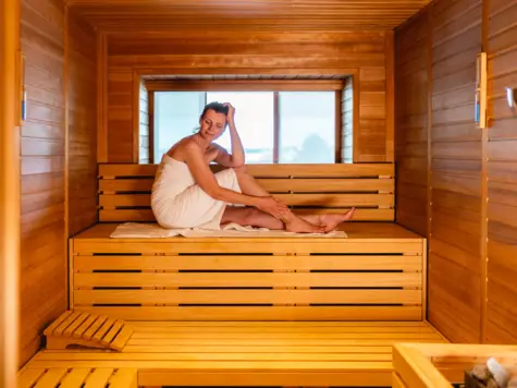  Das Bild zeigt eine entspannte Frau, die in einer traditionellen Holzsauna sitzt. Sie ist in ein Handtuch gewickelt und sitzt entspannt auf den hölzernen Sitzbänken der Sauna. Ihre Haltung und der entspannte Ausdruck ihres Gesichts vermitteln ein Gefühl der Ruhe und Entspannung. Das natürliche Holz der Sauna und das warme Licht, das durch das Fenster einfällt, schaffen eine beruhigende und einladende Atmosphäre. Dieses Bild fängt die essentielle Erfahrung einer Sauna perfekt ein – Entspannung und Wohlbefinden in einer warmen, ruhigen Umgebung.
