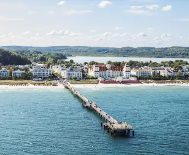 Blick auf ein elegantes Seebad mit klassischer Bäderarchitektur, einer langen Seebrücke und feinem Sandstrand. Im Hintergrund erstreckt sich eine grüne Hügellandschaft unter klarem, blauem Himmel.