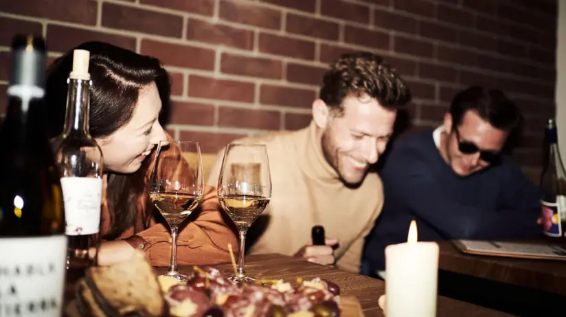 Eine Gruppe von Personen vergnügt sich in einer Bar bei Wein und einem Salami Oliven Board und lacht. 