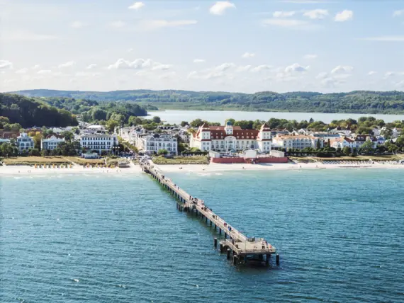 Ein langer Steg führt zu einem Strand vor einer Reihe an Gebäuden mit klarem Himmel und ruhigem Gewässer.