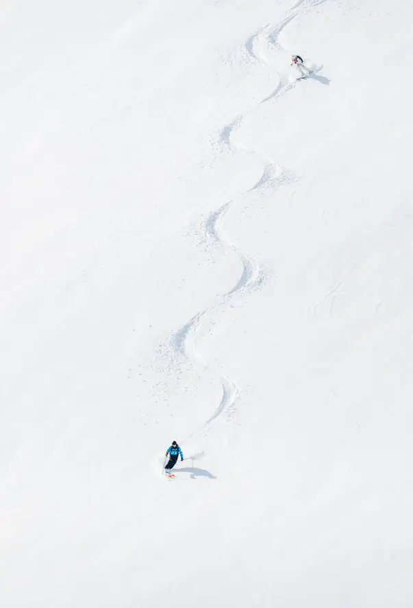 Zwei Skifahrer ziehen ihre Linien durch den Tiefschnee einen Hang hinter.