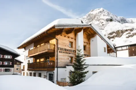 Außenansicht der verschneiten Thurnher’s Residences in Zürs am Arlberg vor alpiner Bergkulisse bei Sonnenschein – ein stilvoller Rückzugsort für den Winterurlaub.