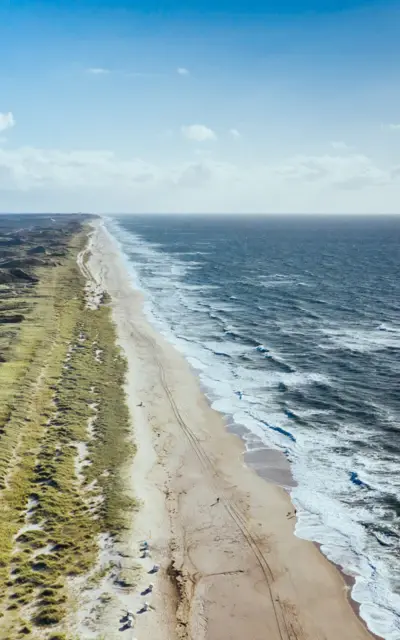 Eine Luftaufnahme vom Strand mit dem angrenzenden welligen Meer und der grünen Dünenlandschaft. 