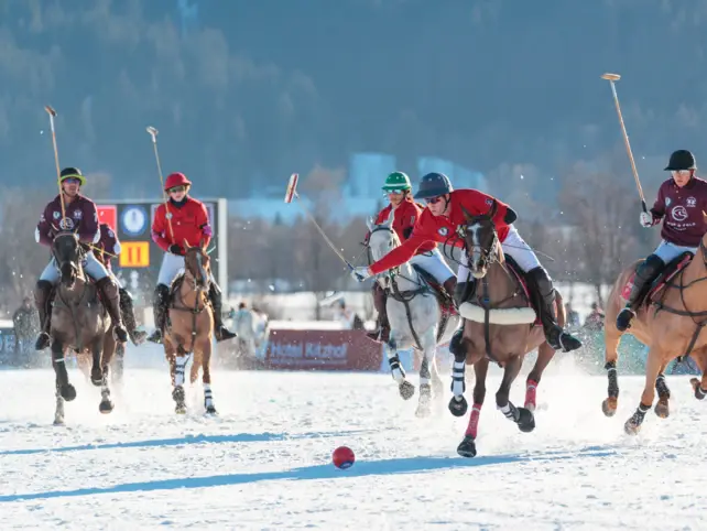 Ein Polospiel auf schneebedecktem Spielfeld mit mehreren Reitern auf ihren Pferden, die mit ihren Schlägern einen roten Ball verfolgen. 