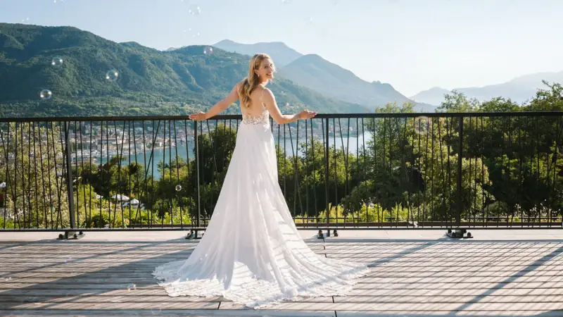 Eine Frau in einem weißen Hochzeitskleid steht auf einer Terrasse mit Geländer und Bergen sowie dem Gardasee im Hintergrund.