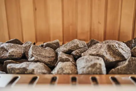  A carefully arranged pile of sauna stones lies ready to store and distribute the heat at the heart of a traditional sauna. The textured gray stones form a natural contrast to the warm wooden interior in the background and embody the authentic sauna experience. 