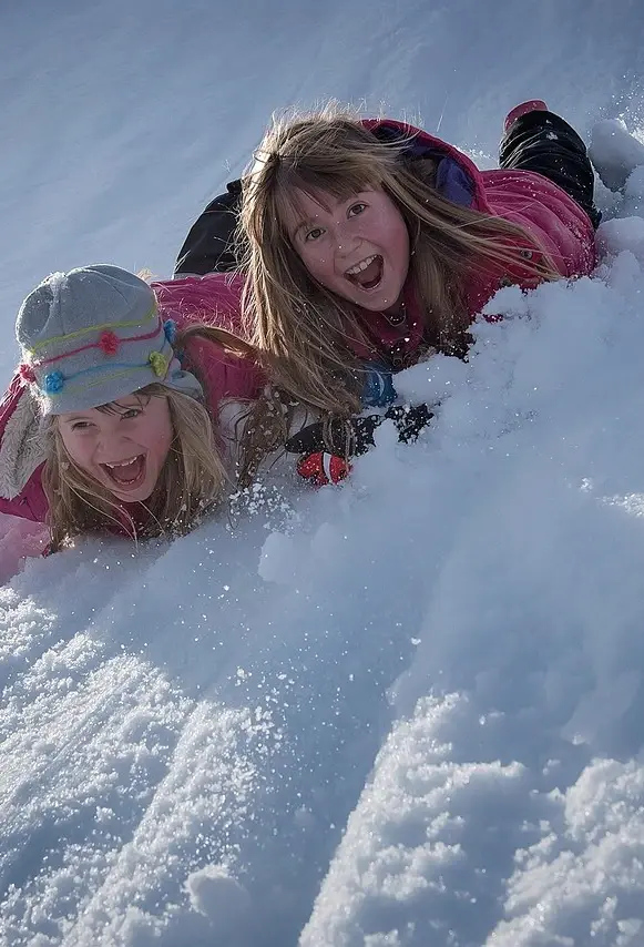 Zwei Mädchen rutschen auf dem Bauch einen schneebedeckten Abhang hinunter und lachen und Kreischen dabei.