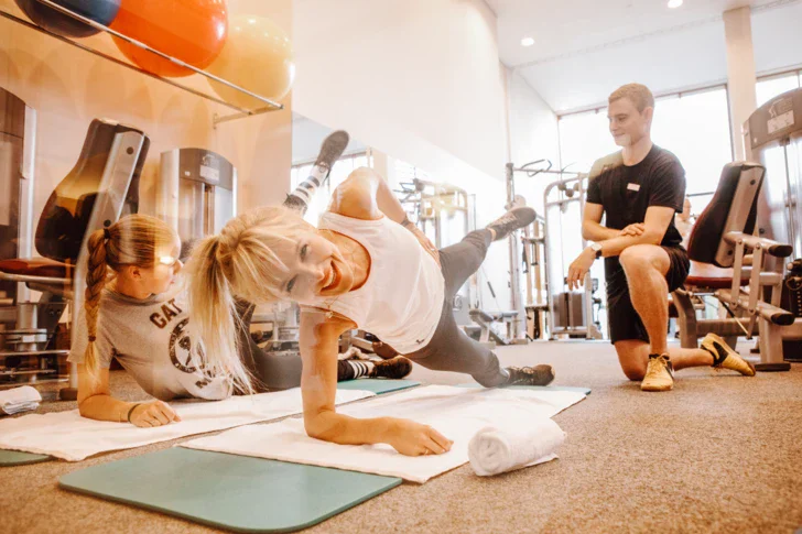 Zwei Frauen trainieren lachend in einem Trainingsbereich mit einem knienden Personaltrainer. 
