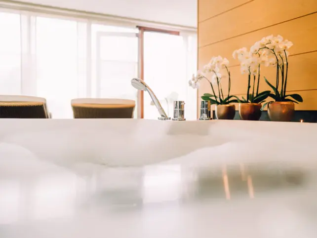 Aus einer mit Wasser und Schaum gefüllten Badewanne wird eine SPA-Suite fotografiert. Zwei Liegen und mehrere weiße Orchideen und eine große Fensterfront sind im Hintergrund zu sehen. 