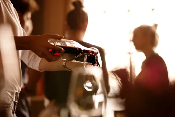 Una mano versa del vino rosso da una caraffa di vetro in un bicchiere da vino e sullo sfondo si riconoscono due donne sfuocate, l'una di fronte all'altra