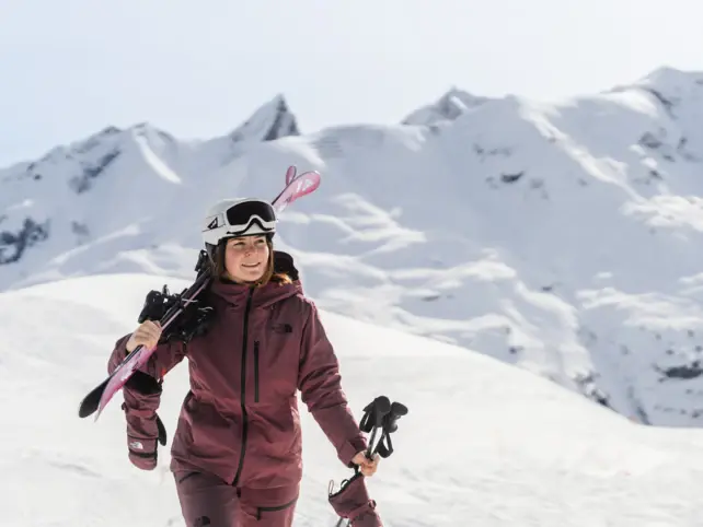 Frau in Skianzug und Helm trägt Skier auf der Schulter und hält Skistöcke in der Hand.