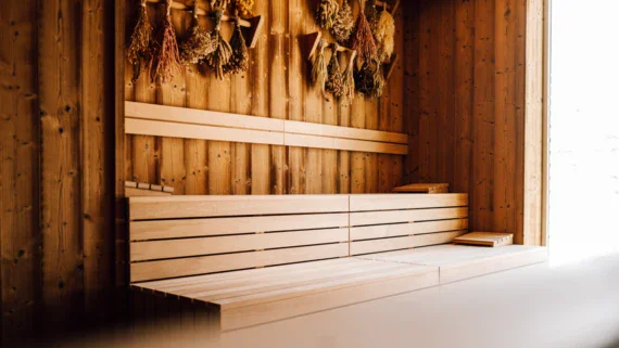 Eine holzverkleidete Sauna mit Stufenbänken. Durch ein Fenster zur rechten Seite scheint warmes, helles Tageslicht herein. An der Wand hängen Bündel von getrockneten Kräutern. 