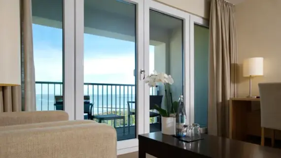 Von einem hellen Sessel wurde die Fensterfront eines Hotelzimmer fotografiert. Im Vordergrund ist ein dunkler Couchtisch zu sehen, auf dem eine Flasche Wasser, zwei Gläser und eine Orchidee stehen. Im Hintergrund ist ein Balkon mit zwei Stühlen und einem Beistelltisch zu sehen.