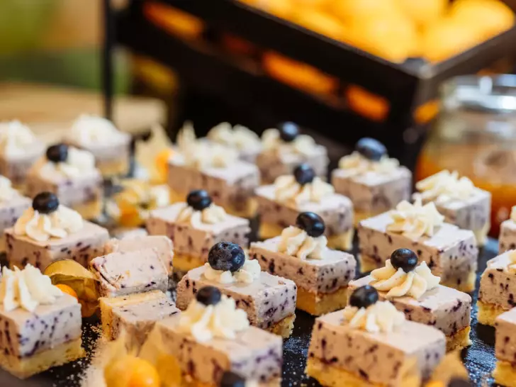 Ein Buffet präsentiert appetitlich angerichtete kleine Kuchenstücke, die mit Creme und frischen Blaubeeren verziert sind. Jedes Stück ist auf einem schwarzen Untergrund platziert und mit Puderzucker bestreut. Im Hintergrund ist ein Korb mit frischen Orangen zu sehen.