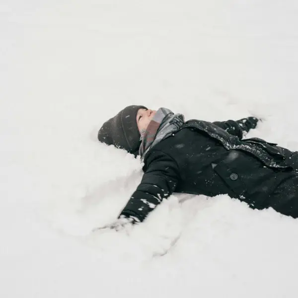 A little boy in a black jacket lies on his back in the snow and makes a snow angel.