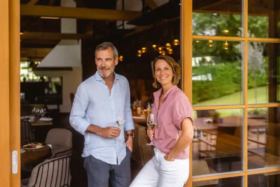 Ein lächelndes Paar steht in der Tür eines gemütlichen, eleganten Restaurants, jeweils mit einem Glas Weißwein in der Hand.