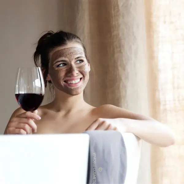 Una donna che indossa una maschera facciale sorride mentre tiene in mano un bicchiere di vino rosso e siede rilassata nella vasca da bagno. Il suo sorriso e il piacere del vino suggeriscono un momento di relax e benessere personale.