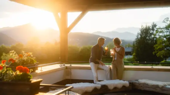 Ein Mann und eine Frau trinken Wein im Freien, umgeben von Pflanzen vor der Kulisse eines Sonnenuntergangs in den Bergen..