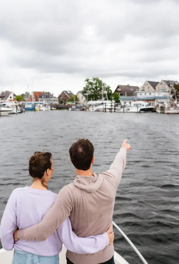 Eine Frau und ein Mann stehen am Bug auf einem Boot und blicken auf einen kleinen Hafen. Der Mann streckt dabei seinen rechten Arm aus und zeigt auf die Boote im Hafen.