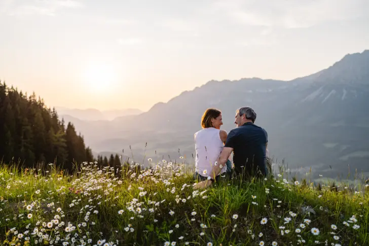 Ein Mann und eine Frau sitzen auf einem blumenübersäten Hügel mit Bergkulisse im Hintergrund.