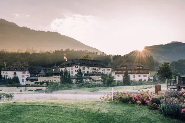 Ein malerisches Hotel in den Bergen, umgeben von grünen Wiesen und blühenden Gärten. Die Sonne geht hinter den Bergen auf, und der Morgennebel steigt über einem kleinen See auf.