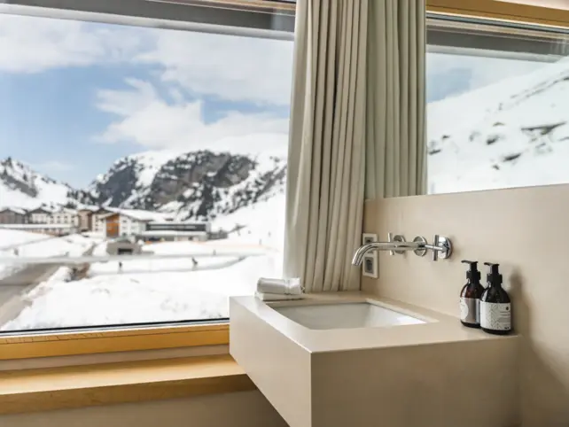 Ein beiges Badezimmer mit einem großem Waschbecken, was vor einem großem Fenster mit Ausblick auf die verschneite Bergwelt steht.