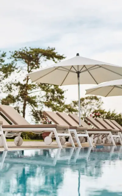 Poolliegen aufgereiht an einem Swimmingpool im Freien unter weißen Sonnenschirmen mit Bäumen im Hintergrund.