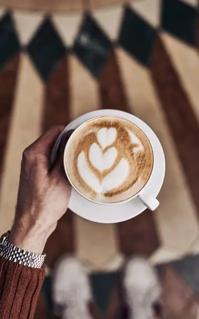 Person hält eine Tasse Cappuccino über einem gemusterten Fliesenboden.