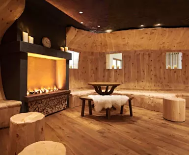 Ein gemütlicher Raum mit holzverkleideten Wänden und Boden, ausgestattet mit einem Kamin, Kerzen, einem rustikalen Holztisch, Hockern und einer Bank mit einem flauschigen Lammfell. Das Ambiente ist warm und einladend mit indirekter Beleuchtung.