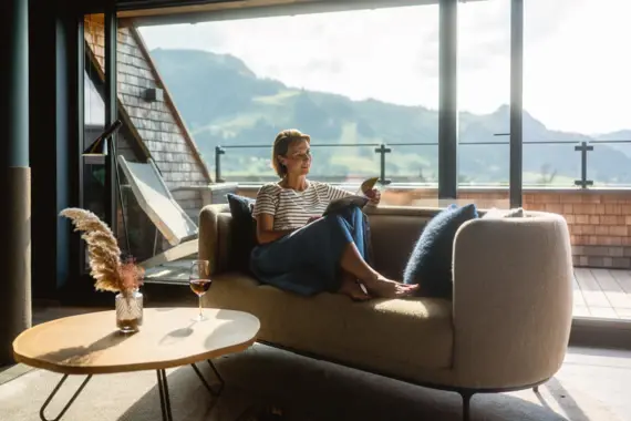 Eine Frau sitzt auf einem Sofa und liest ein Buch. Im Hintergrund ist durch ein großes Fenstern eine grüne Bergkulisse zu sehen. 