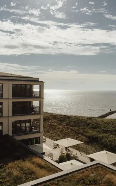  Ein modernes Hotelgebäude mit Balkonen, Sonnenschirmen und Meerblick, die Sonne glitzert auf dem Wasser.