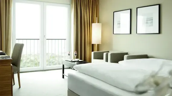 A hotel room with a bed in the foreground and a desk and chair on the left. At the back right of the room are two light-colored armchairs, a floor lamp and a coffee table with two champagne glasses filled with fruit.