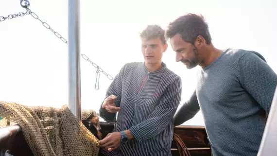 Zwei Personen an Bord eines Fischerbootes bei der Arbeit, sichtbar vertieft in die Überprüfung der Fischernetze. Das Tageslicht sorgt für eine helle und freundliche Atmosphäre. Die Szene strahlt die Authentizität des Fischerlebens aus, mit einem Fokus auf Handarbeit und Teamarbeit.
