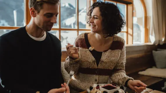 Ein glückliches Paar genießt ein Dessert in einem gemütlichen Restaurant mit großen Fenstern, die den Blick auf eine verschneite Berglandschaft freigeben. Die Frau lacht, während sie mit einer Gabel etwas vom Teller nimmt, der mit Beeren und einer Soße dekoriert ist.