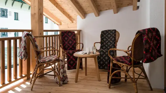 Auf einem Balkon aus hellem Holz stehen vier Stühle mit jeweils einer Decke über der Rückenlehne. In der Mitte der Stühle steht ein kleiner Holztisch mit einem Buch und zwei Getränken.