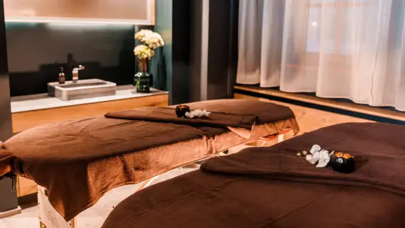 Two massage tables, each with brown towels, a small candle and orchid flowers. A washbasin and a vase of flowers can be seen in the background.