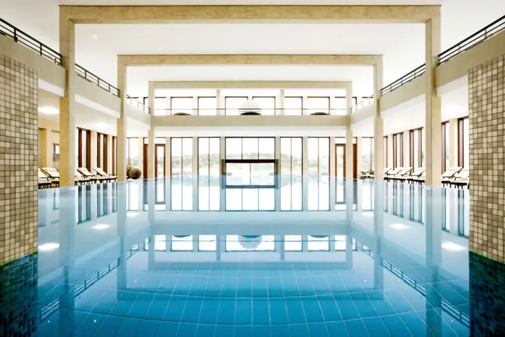 Ein eleganter Poolbereich mit großen, bodentiefen Fenstern durch die das Tageslicht den Raum durchflutet. Zu den Seiten des Beckens stehen hohe, bis zur Decke reichende Säulen und helle Liegen bieten Möglichkeit zur Entspannung. 