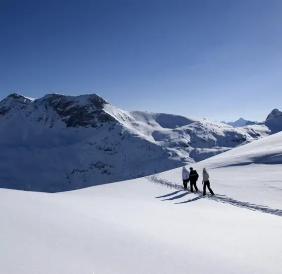 Three people walk through a snowy landscape with mountains in the background.