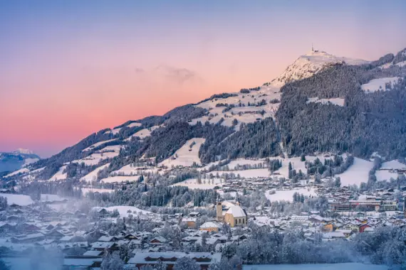 Schneebedeckte Berge sind von der aufgehenden Sonne in warmes Pink getaucht. Im Tal der Berge liegt ein kleines Dorf mit Kirche. 