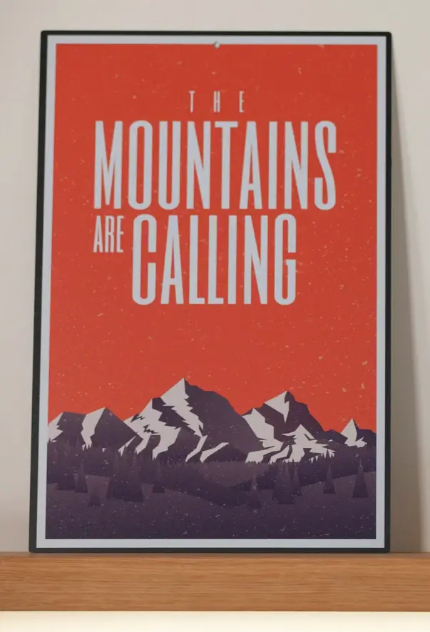 A picture in a picture frame stands on a wooden shelf. The illustration shows a mountain range with a red background bearing the words "The mountains are calling".