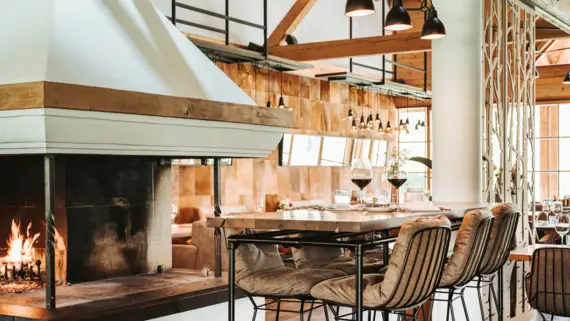 Ein gemütliches Restaurant mit modernem alpinen Design: Im Vordergrund steht ein offener Kamin mit loderndem Feuer, daneben eine stilvolle Bar mit hohen Stühlen. Im Hintergrund sind elegant gedeckte Tische und warme Beleuchtung zu sehen, die eine einladende Atmosphäre schaffen.