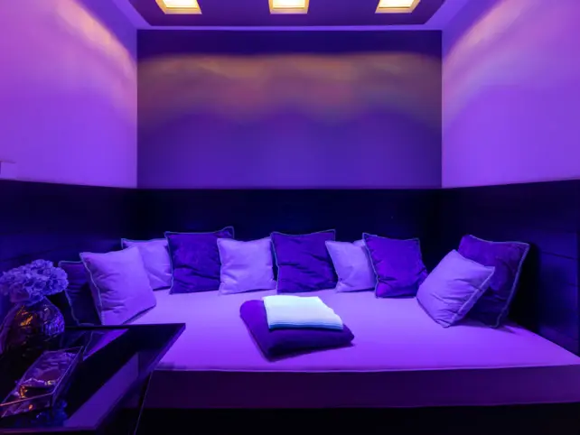 Ein lilafarben beleuchteter Entspannungsbereich mit einer großen Liegemöglichkeit und vielen gemütlichen Kissen 