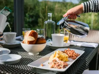 Ein Frühstückstisch mit Lachs und Rührei auf einem Teller. Daneben steht ein Brötchenkorb und eine Hand gießt Kaffee in eine Tasse. Der Hintergrund zeigt grüne Vegetation. 
